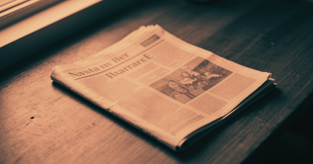 A single newspaper folded on a bare wooden table, masthead visible, strong window light from one side