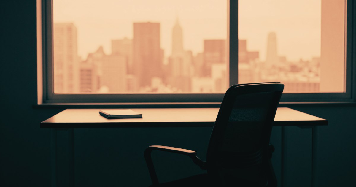 A single chair at an empty desk facing a large window overlooking a city skyline