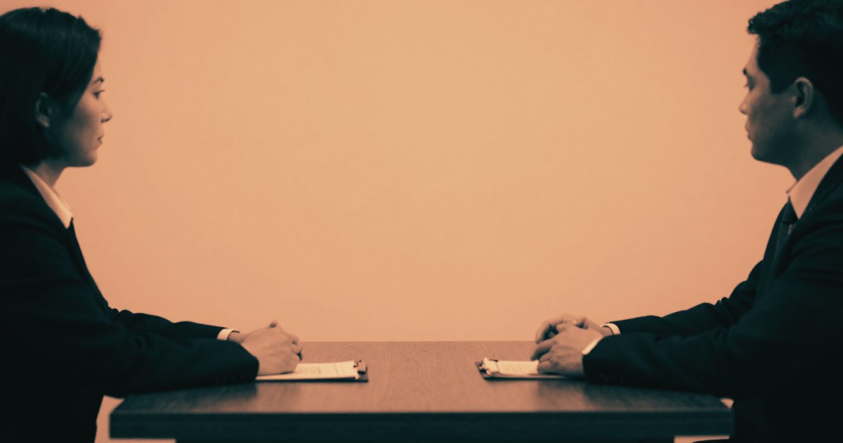 Two people seated across a table, shot from the side at distance, faces not visible, one person's hands flat on the table