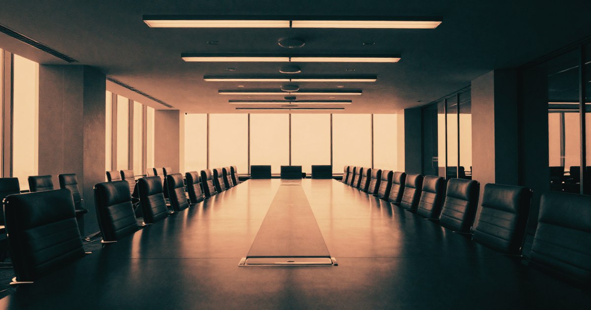 A vast empty boardroom shot from the end of a long table, chairs in place, no one seated